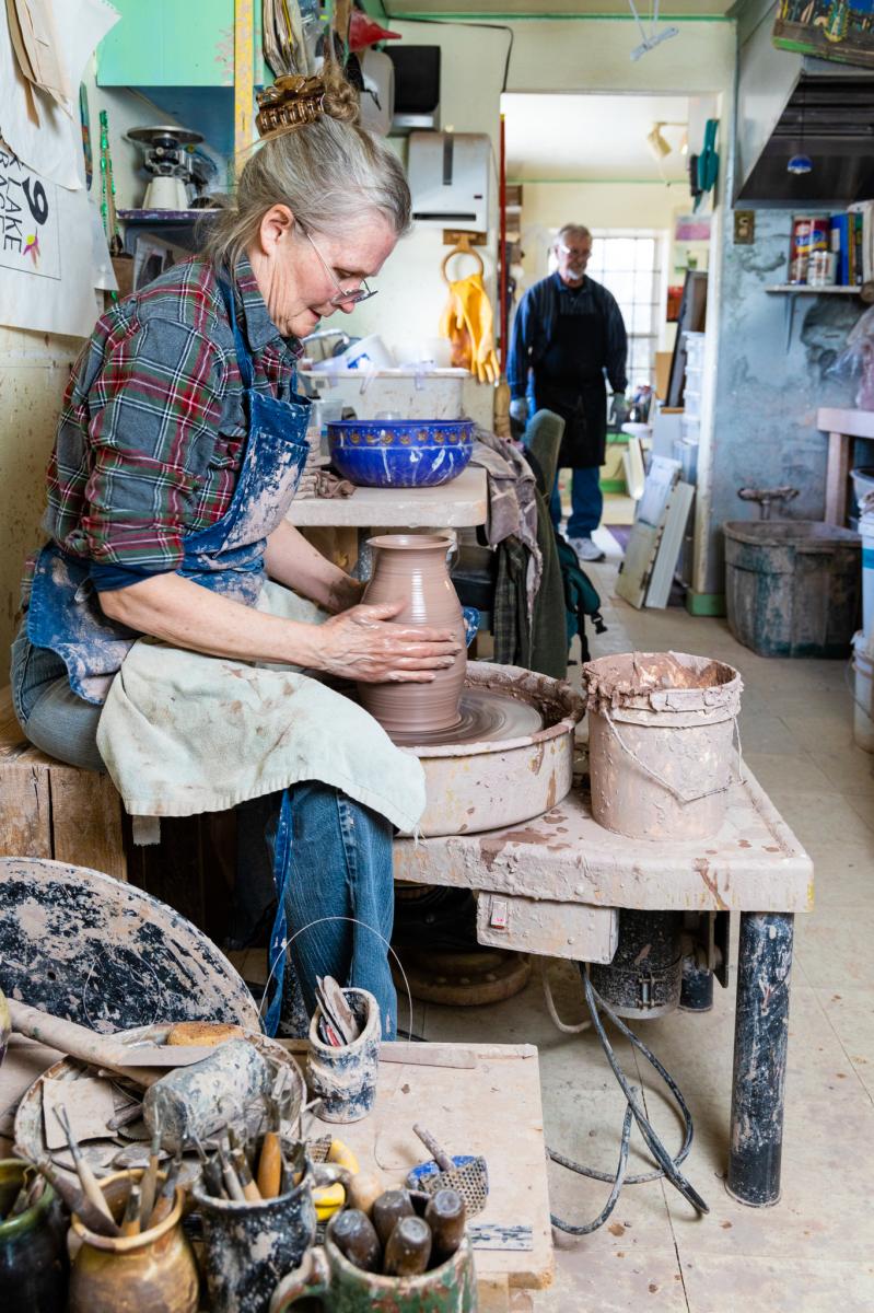 A person shapes a pot on a pottery wheel in a cluttered workshop, with another individual observing from the background.