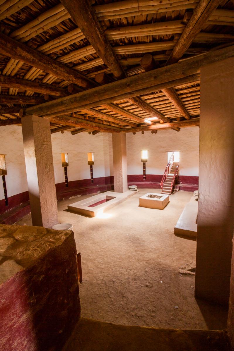 Interior view of a room in Aztec Ruins National Monument featuring traditional Pueblo architecture with wooden beams, large stone pillars, and minimalistic furnishings.