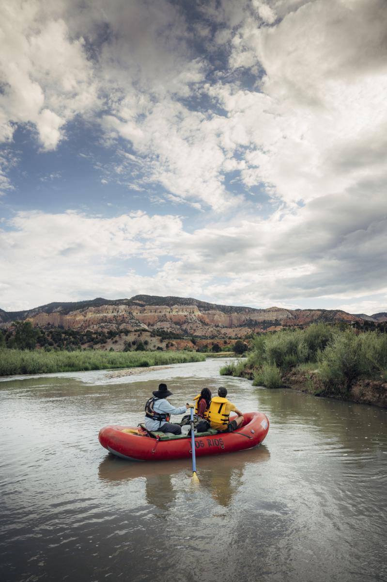Three people rafting on a river with lush greenery and a scenic, rocky backdrop under a cloudy sky.