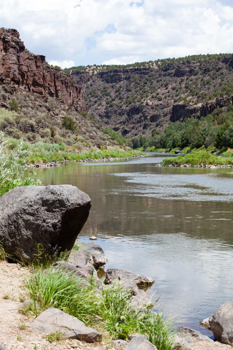 View of the Rio Grande flowing through a scenic canyon with rocky shores and lush greenery under a partly cloudy sky.