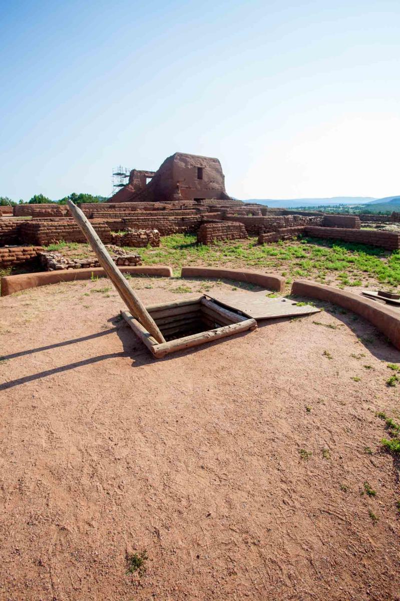 View of Pecos National Historical Park with the hatch entrance to a kiva in the foreground and the ruins of a large adobe structure in the background under a clear blue sky.