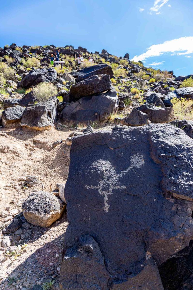 Petroglyphs on volcanic rock at Petroglyph National Monument, with a visitor examining them in the distance under a clear sky.