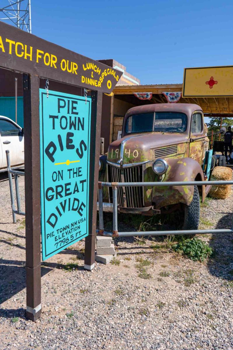 Sign reading "Pie Town Pies" next to an old, rusty pickup truck in front of a colorful establishment with signs advertising lunch and dinner options.