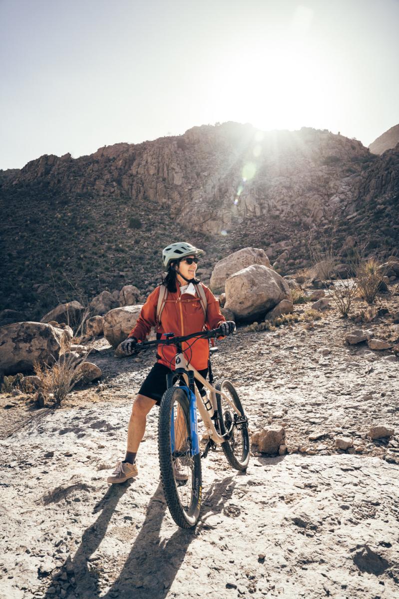 A person with a helmet stands next to a mountain bike on a rocky desert trail, with rugged mountains in the background and sunlight streaming through.
