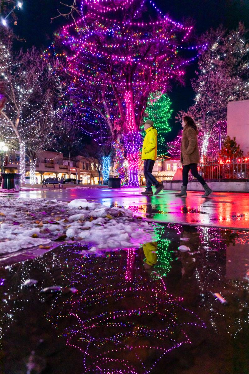 Two people walking by illuminated trees in a festive outdoor setting, with colorful lights reflected on snowy, wet pavement.
