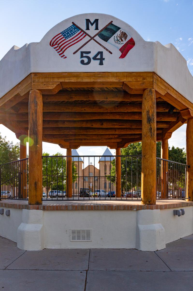 A gazebo with wooden beams supporting a peaked roof featuring paintings of the U.S. and Mexican flags, labeled "M 54", set against a clear blue sky.