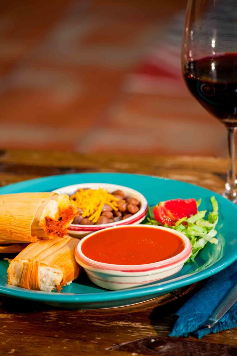 A plate of Mexican tamales accompanied by sides of beans, salad, and a bowl of salsa, served with a glass of red wine.