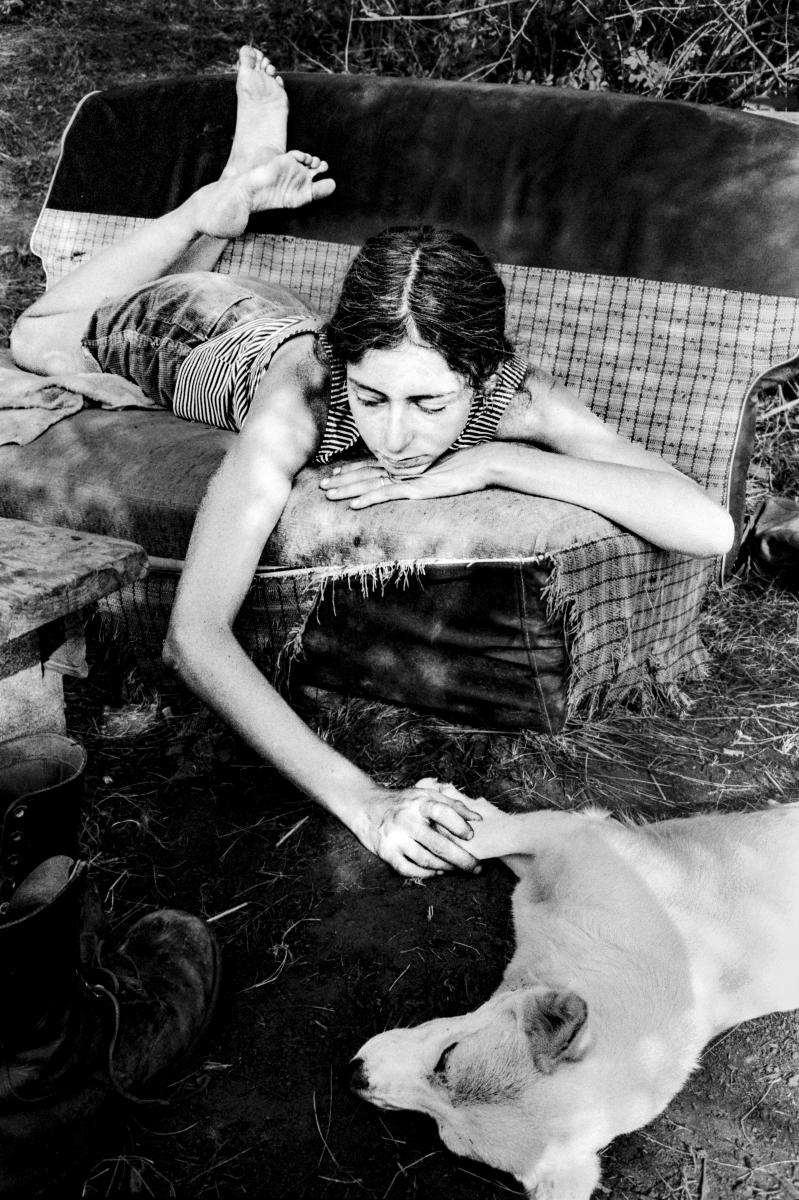 Black and white photograph of a woman lying across a worn outdoor couch and reaching toward a dog resting below her.