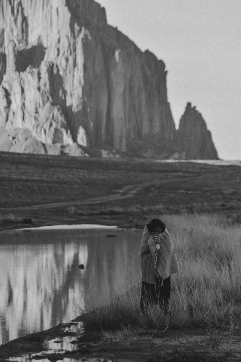 A person wrapped in a blanket stands by a calm river, with tall, rugged cliffs in the background.
