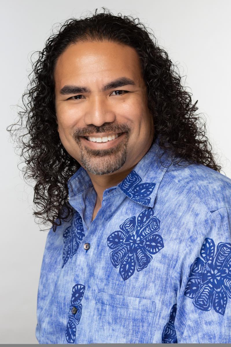 Headshot of a Hawaiian man in a blue collared shirt with long hair smiling at the camera.
