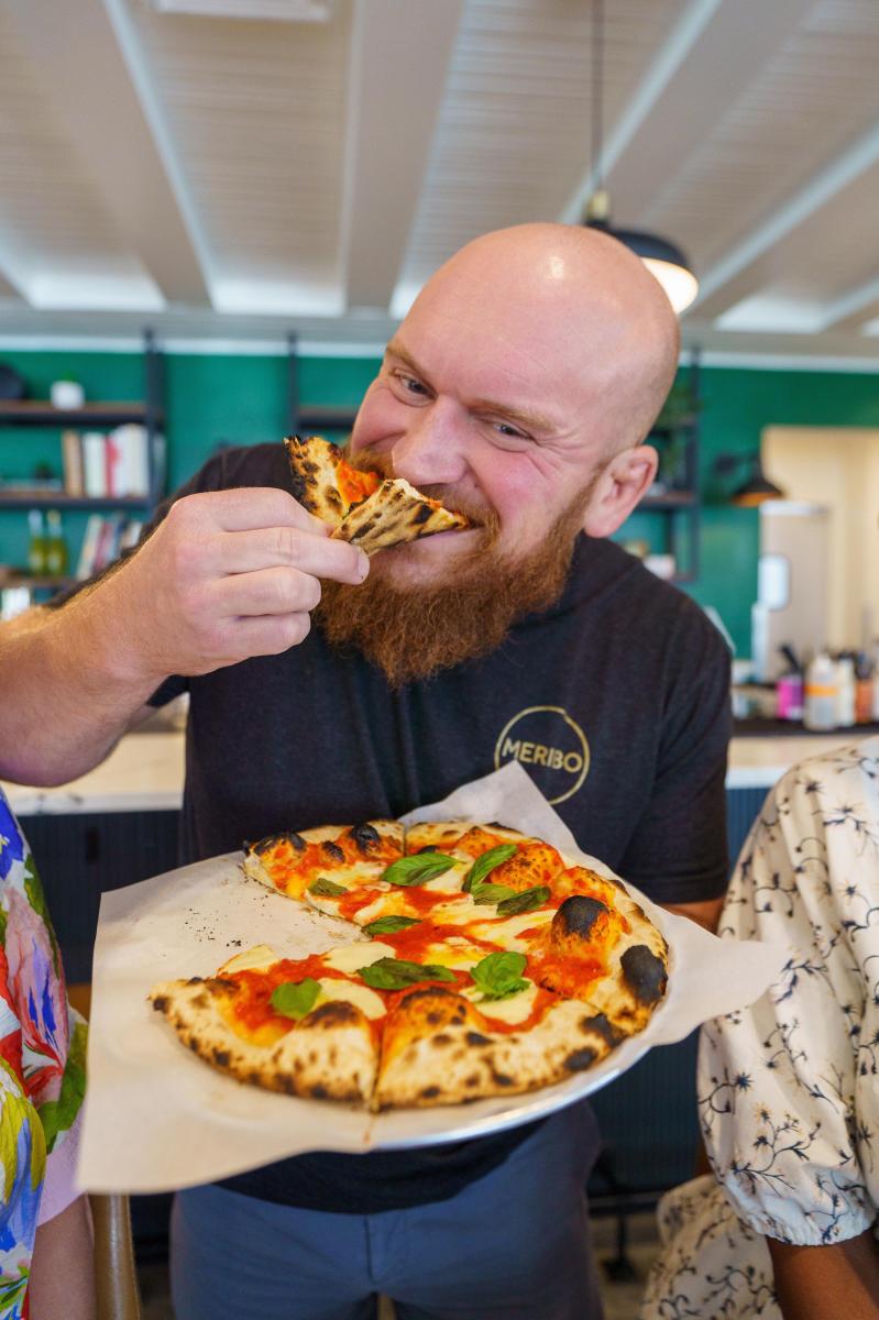 Chef Gavin Jobe of Meribo takes a bite of a wood-fired Margherita pizza while holding the pie inside the restaurant's bright, modern dining room.