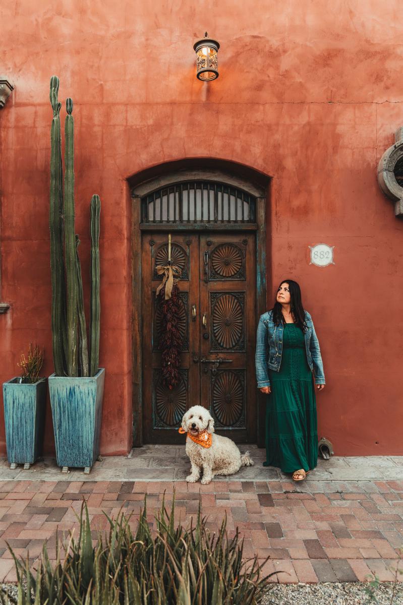 Woman with a dog standing in front of an orange-red Spanish style home with an ornanted wood door and potted cacti