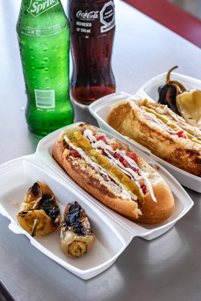 Sonoran hot dog with two peppers in a take out container and sodas on a table