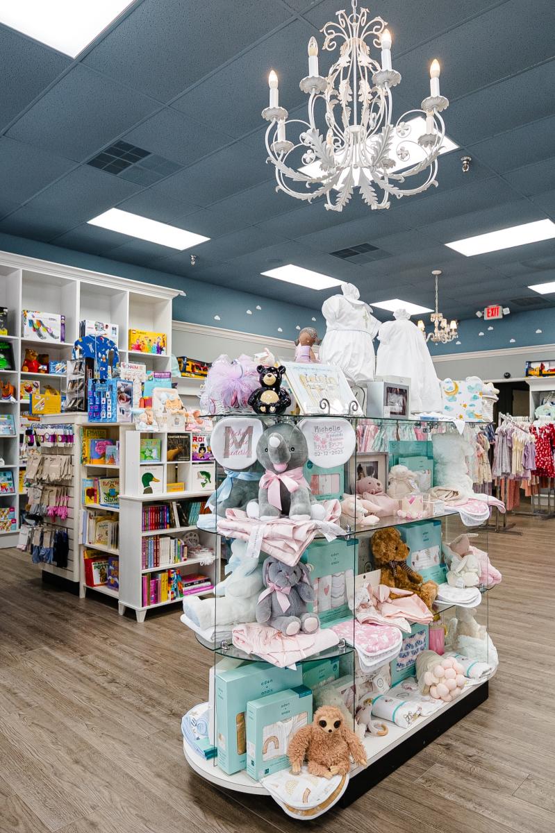 A denim-blue shop with white chandeliers and detailing. A glass display is stocked full of baby plushies, bibs, baptism gowns, and more. In the back are white shelves of baby books and toys.