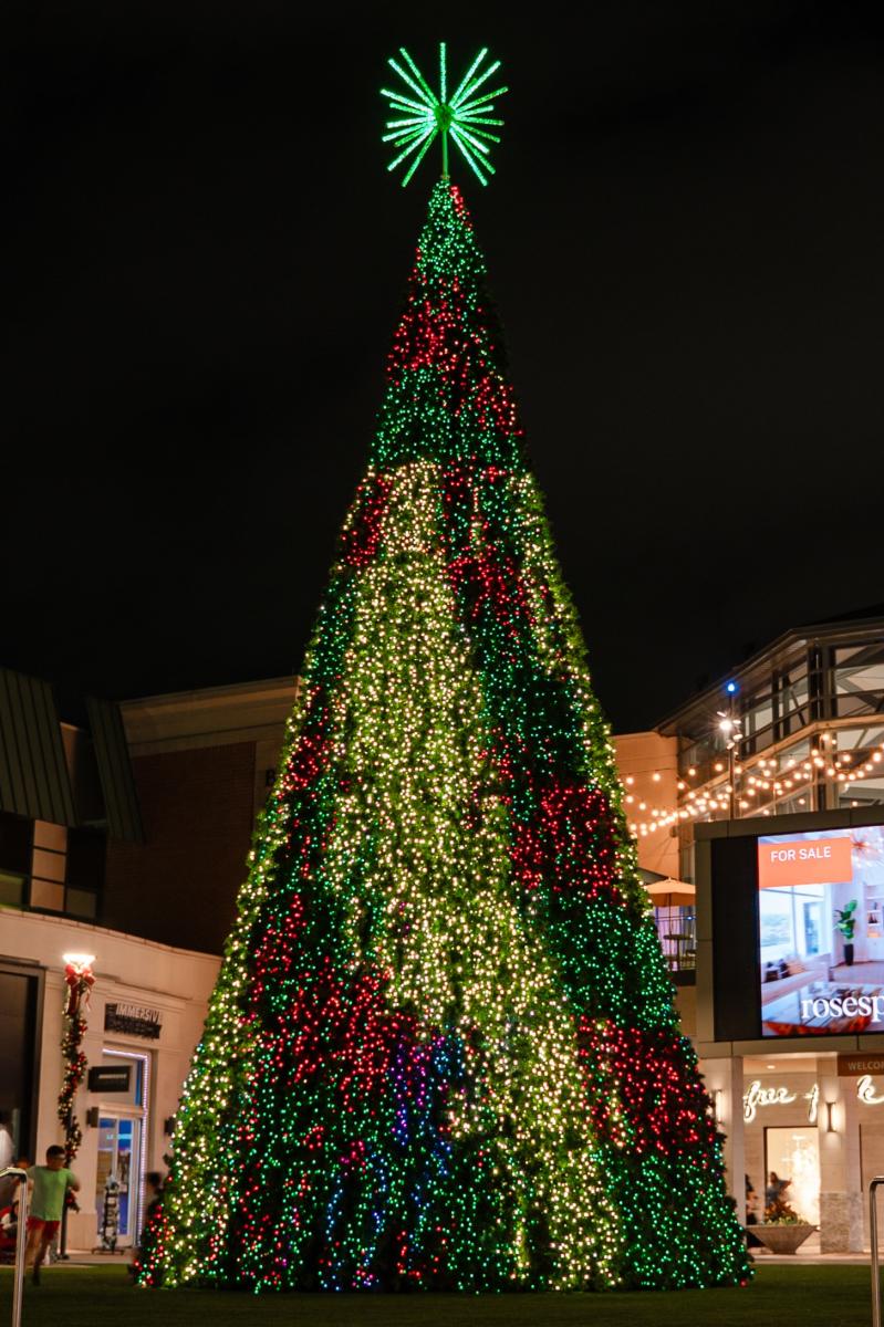 The Christmas Tree at The Mall features a flashing light show, currently transitioning from red and green to gold. A bright green starburst tops the tree.