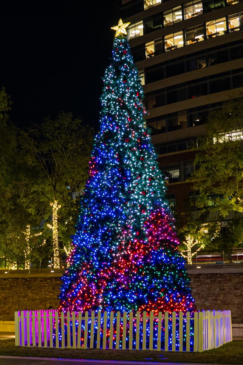 The Christmas Tree at Waterway Square features a flashing light show, currently transitioning from green and red to blue and purple. A bright gold star sits on top. In the background are a brick fountain, trees wrapped in gold twinkle lights, and a tall office building with several lights on.