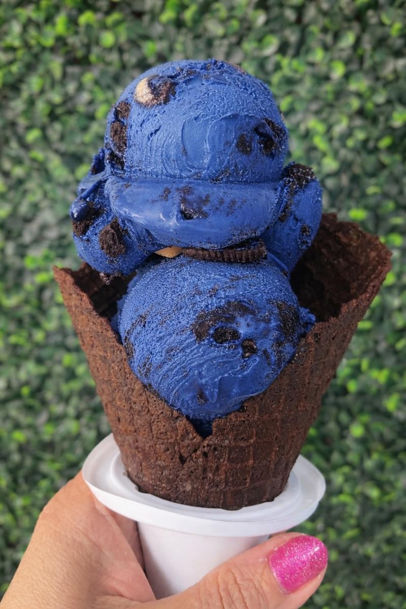 A hand holds up a waffle cone with large scoops of dark blue ice cream with Oreo chunks.