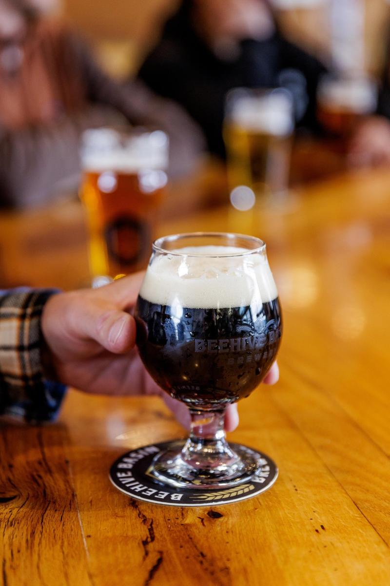 A hand holds a dark beer in a glass on a wooden table, with blurred patrons and various beer glasses in the background.