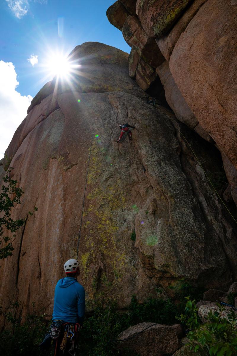 Climber lead climbing on a vertical rock face at Vedauwoo, Wyoming, with a belayer below and the sun illuminating the dramatic rock formations.