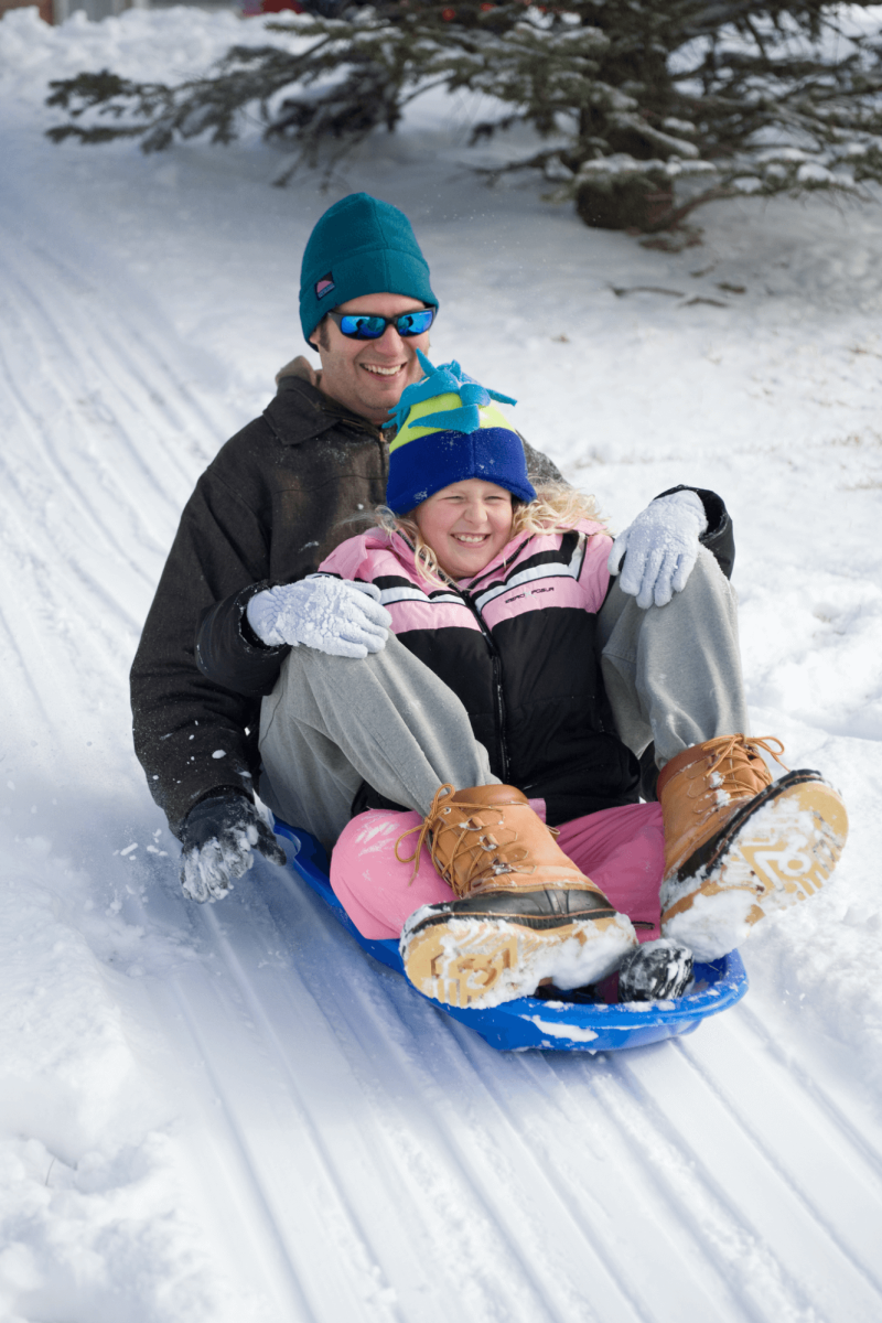 A man and a young girl joyfully sledding down a snowy slope, both wearing winter attire with trees and snow-covered ground in the background.