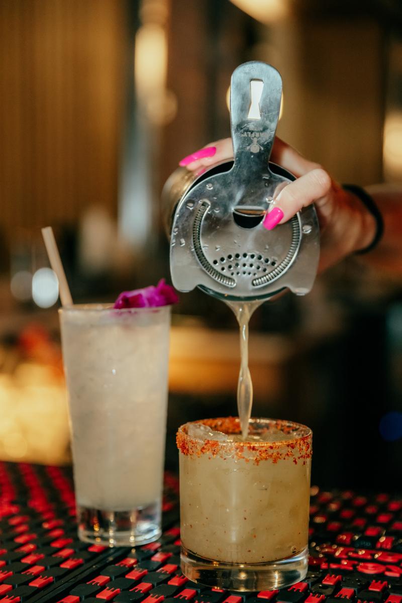 Image is of two cocktails with a hand, pouring liquid into the smaller cocktail glass.