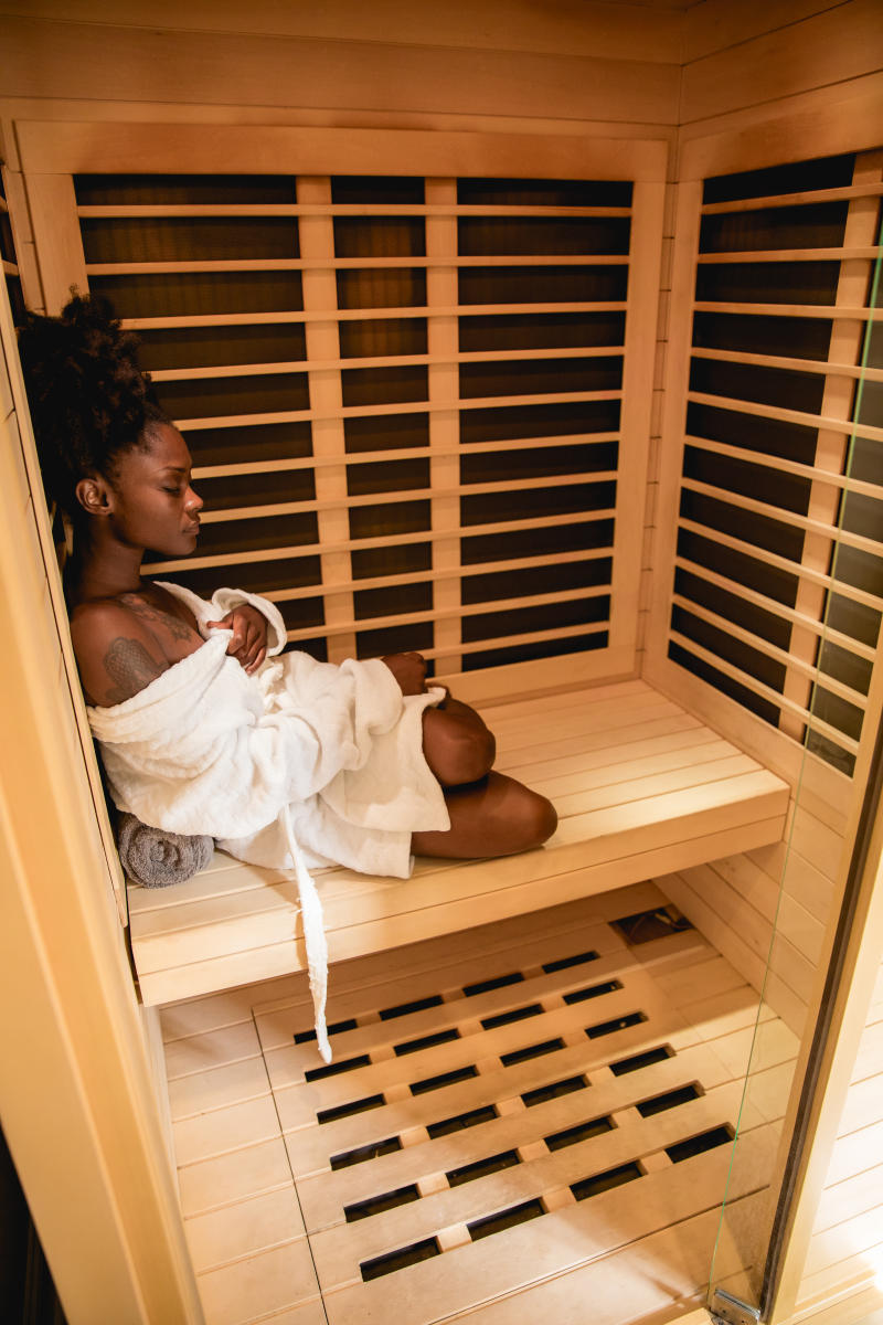 A black woman sits peacefully in a cedar sauna, wrapped in a white robe with eyes closed, enjoying a calming self-care moment in Fayetteville, NC's Floating Shanti Spa.