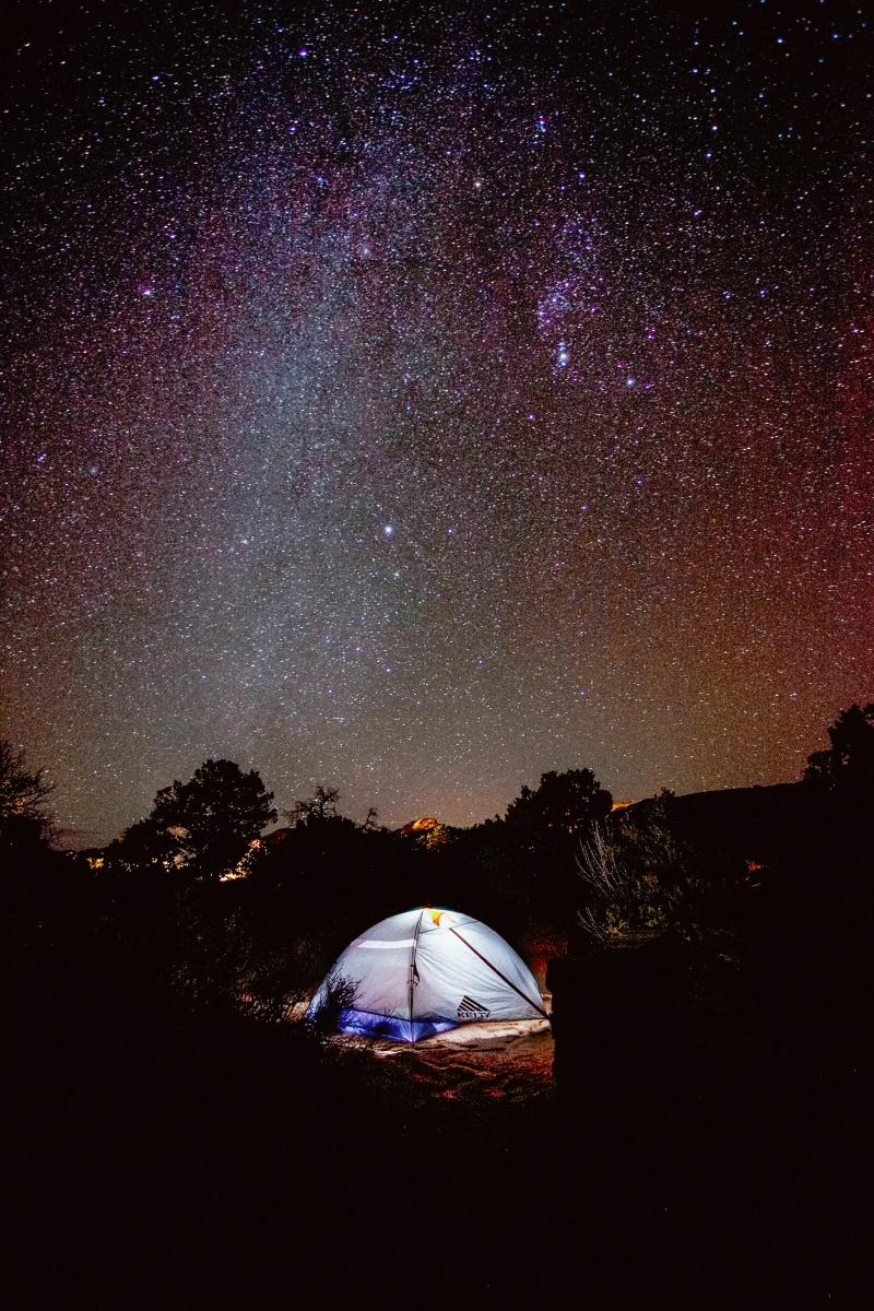 Countless stars and the Milky Way illuminate the dark night sky above a glowing tent at Arches National Park.