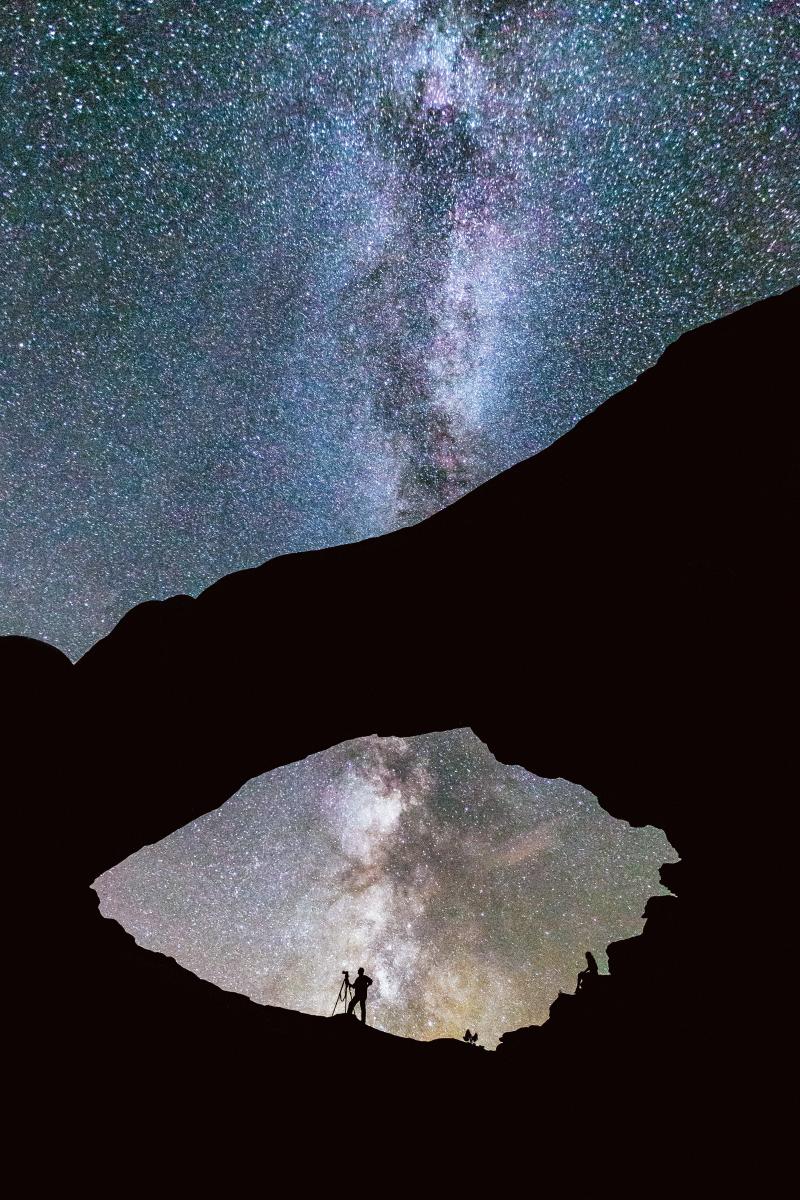 The Milky Way rises above a silhouetted photographer and a canyon rim in Moab's desert landscape at night.