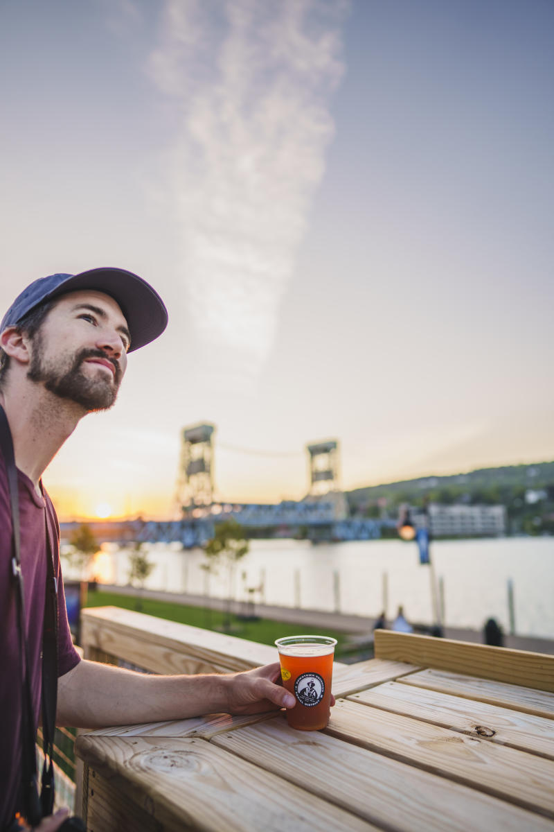a person smiles looking to the distance holding a cup of beer, the lift bridge and waterfront in the background