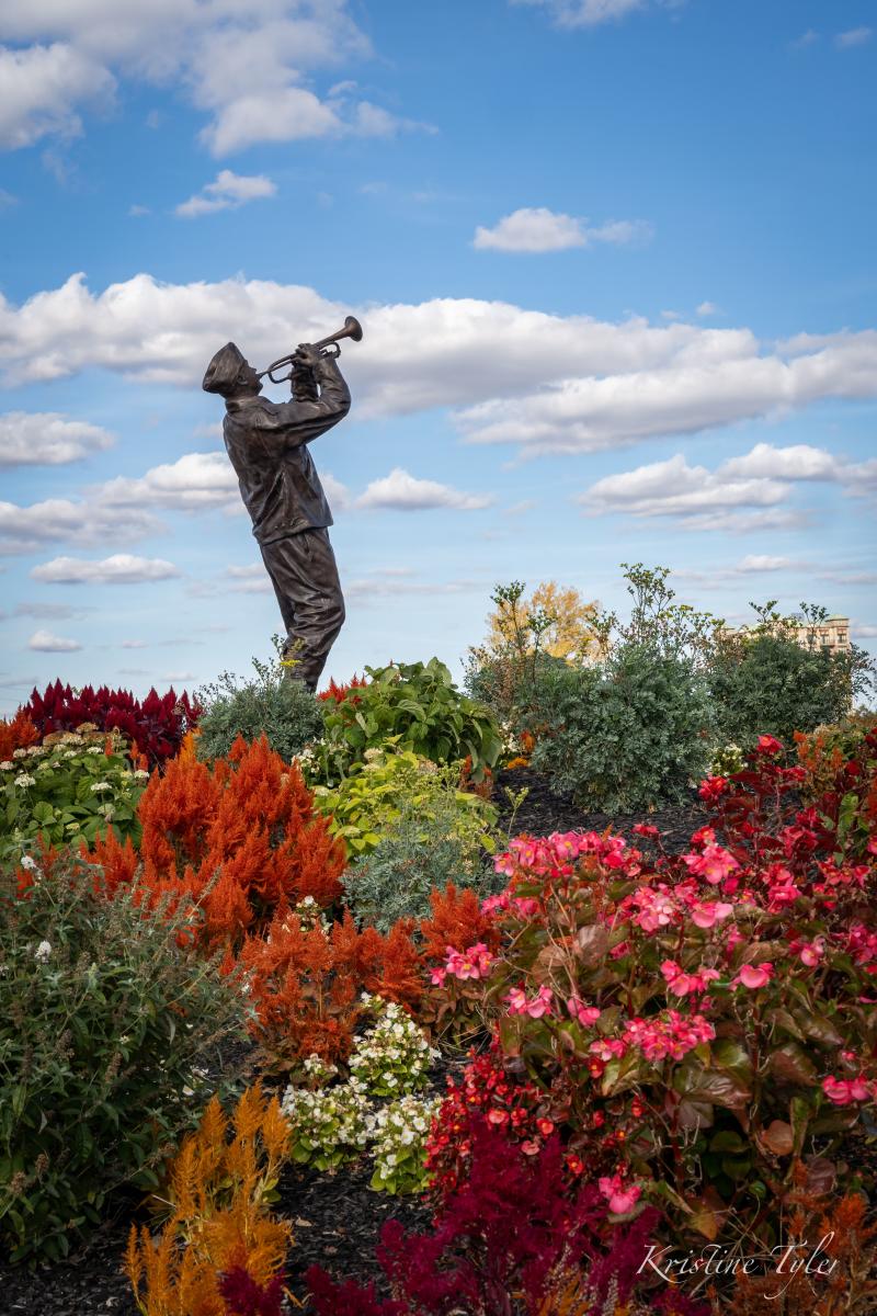 Statue of a person playing a trumpet, set amidst a vibrant garden of multicolored flowers and plants, under a partly cloudy sky.