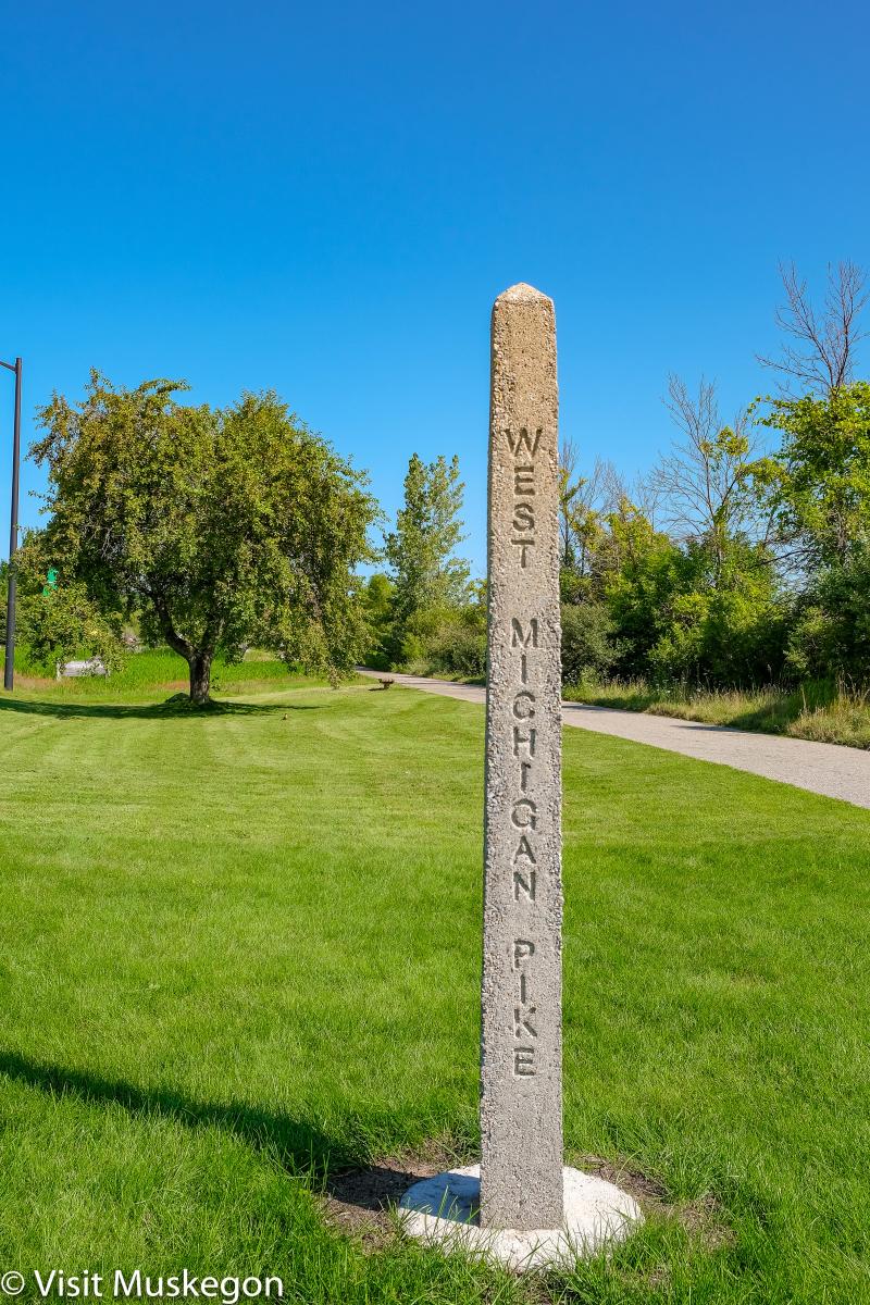 Concrete, pyramidal shaped West Michigan Pike Marker rises above green grass. Blue sky, cement bike path and green trees are in background.