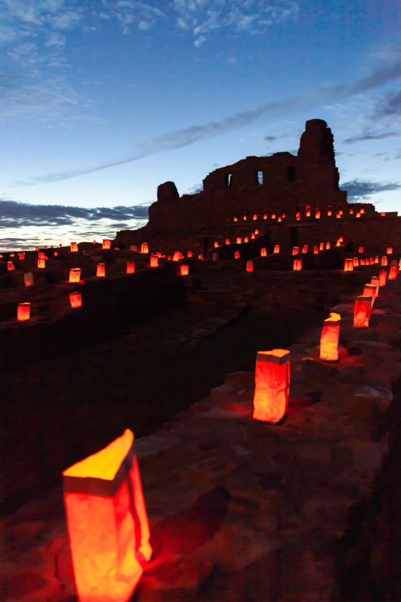Luminarias, or farolitos illuminate a path leading to the ancient footprint of a building against the backdrop of a dusky sky.
