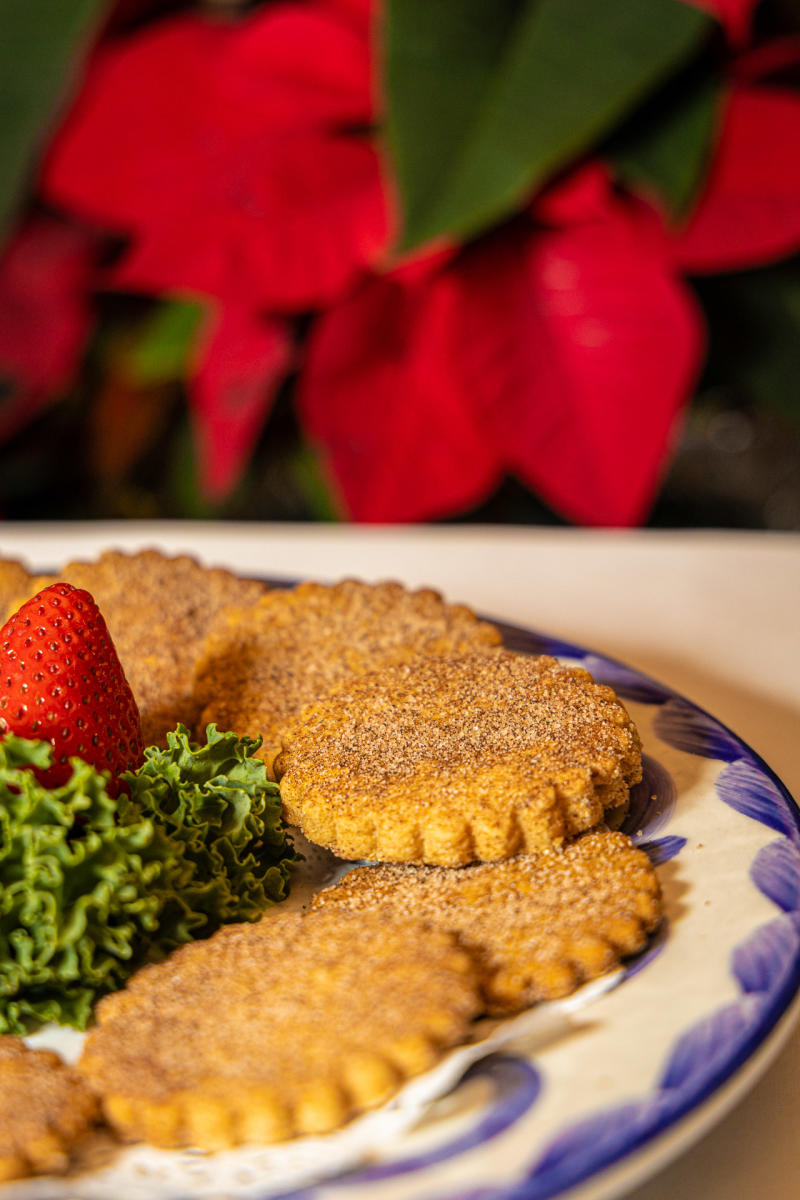 A plate of sugar coated biscochitos arranged with a strawberry and a garnish of curly kale, set against a backdrop of vibrant red flowers.