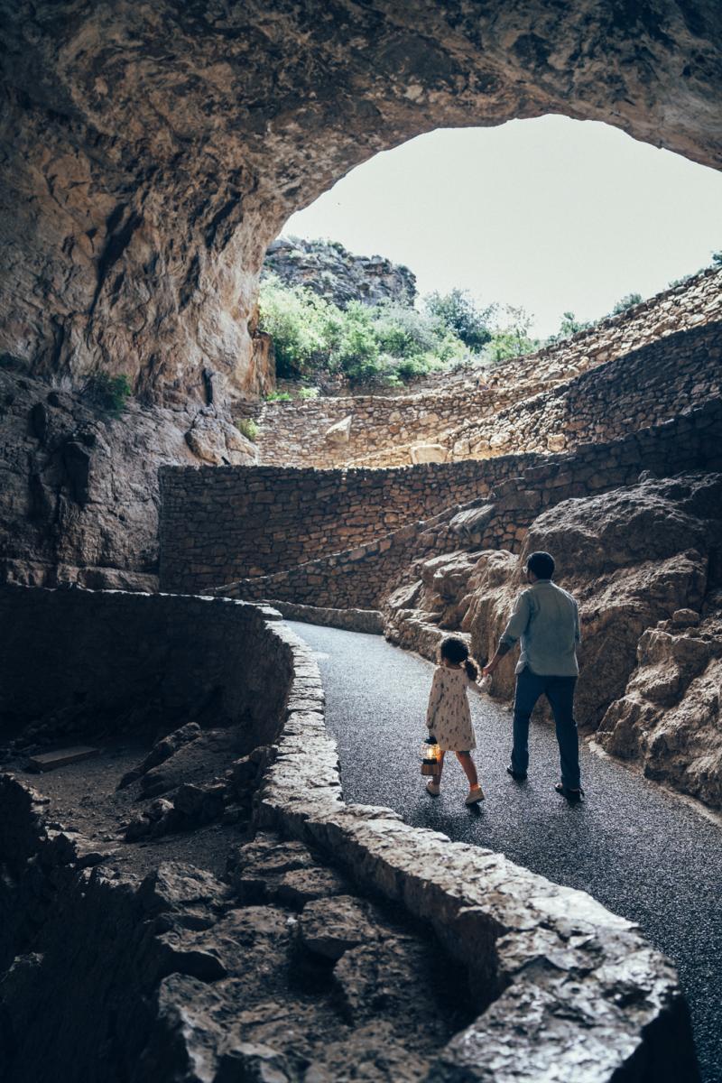 A person and a child are walking hand in hand through Carlsbad Caverns, following a winding pathway that is surrounded by rocky walls and lit by daylight streaming through the cavern's opening.