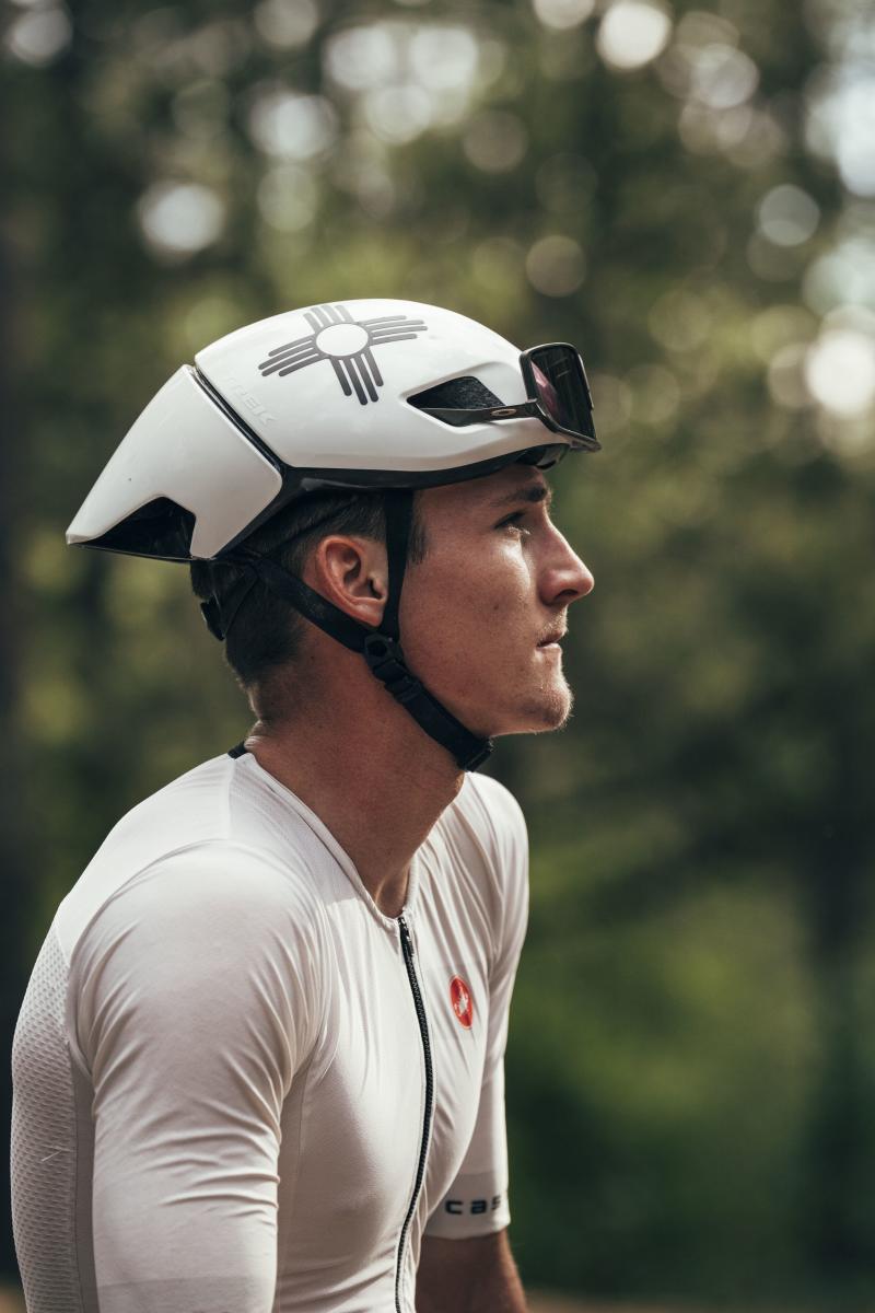 A person wearing a white cycling helmet and jersey is looking to the side. The helmet features a logo resembling the Zia symbol. The background is blurred greenery.