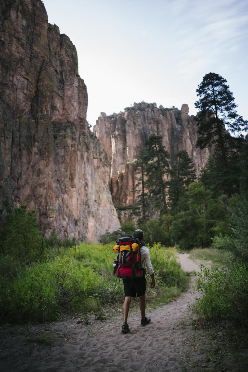 A person with a backpack walking on a trail in a forested area with tall, rocky cliffs in the background.