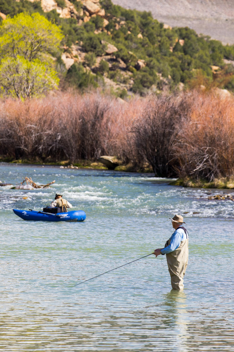 Two people enjoy outdoor activities on a river. One person stands in the foreground fly fishing, while another person in the background maneuvers an inflatable kayak through gentle rapids. The river is surrounded by lush trees and rocky hills.