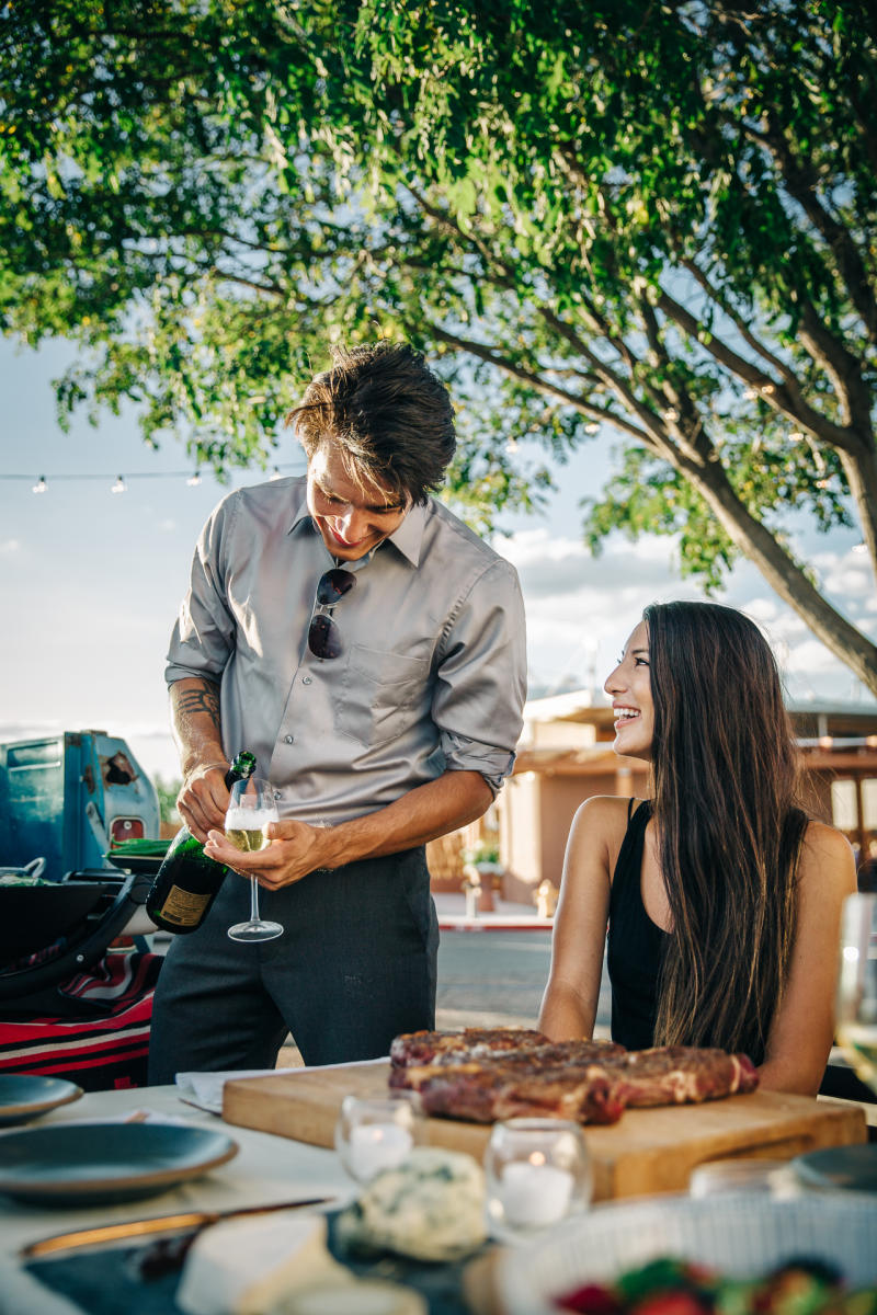 In a sunlit outdoor setting, a person is pouring a drink into a glass held by another person who is seated at a table with cuts of meat. The background features trees and a traditional adobe building.
