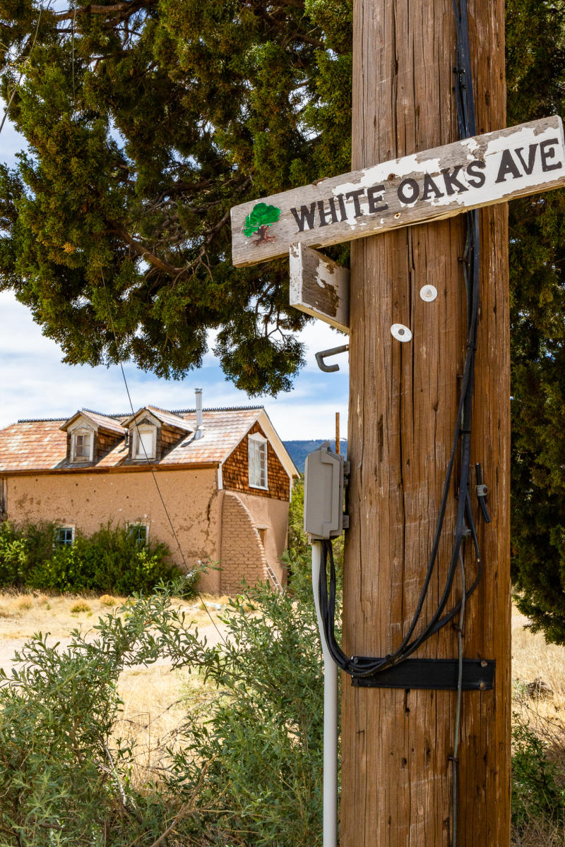 A rustic street scene shows a wooden utility pole with a sign for "White Oaks Ave" amidst greenery. In the background, a historic building with a shingled roof and dormer windows stands under a clear sky.