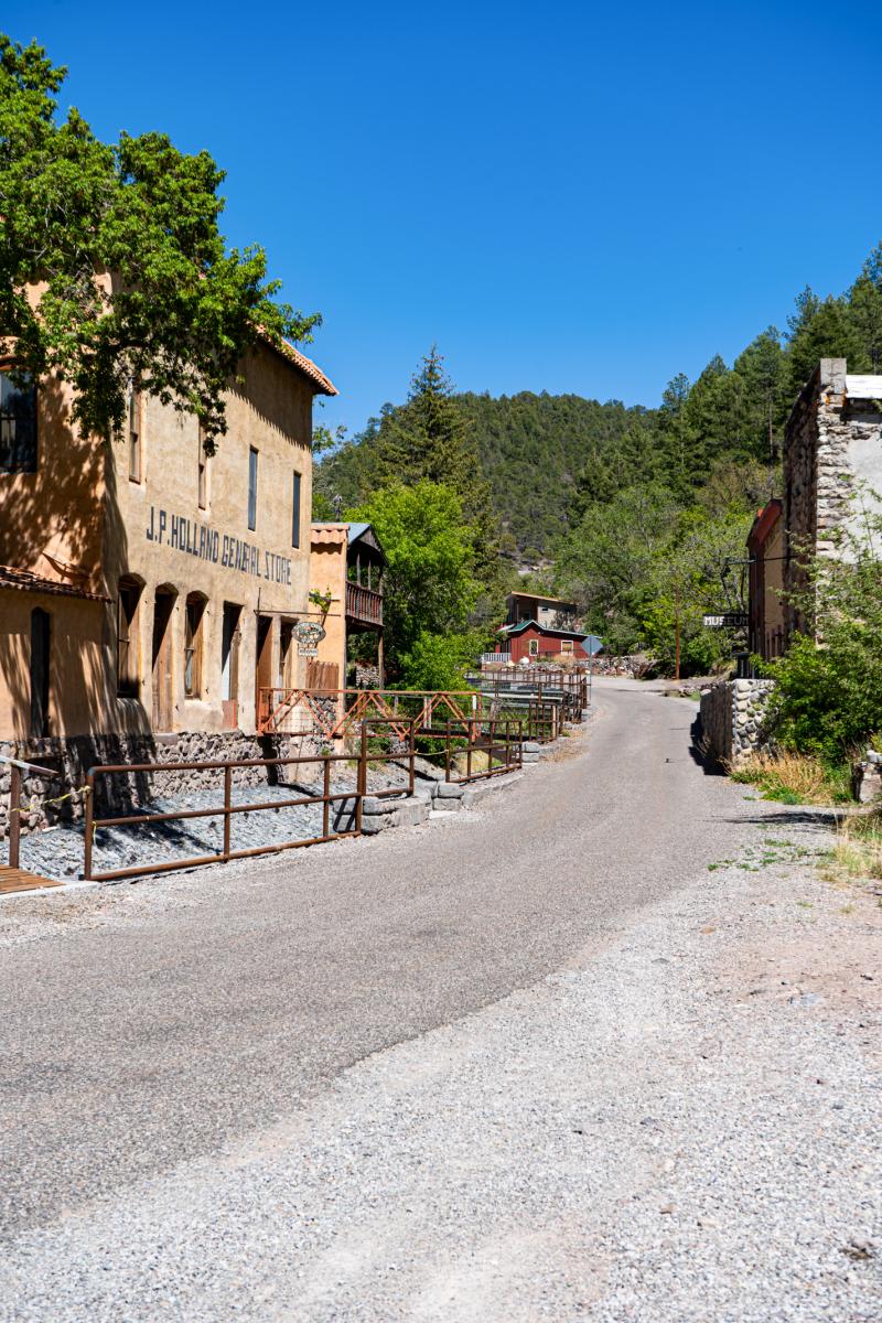 An image of a narrow street running through the historical J.P. Holland General Store in a rustic village, surrounded by lush greenery and mountains in the distance.