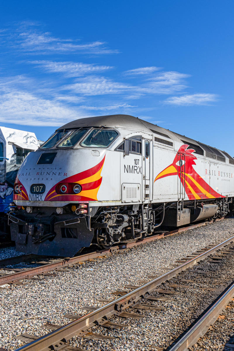 A New Mexico Rail Runner Express train on a track under a clear blue sky. The train features a distinctive, colorful roadrunner design.