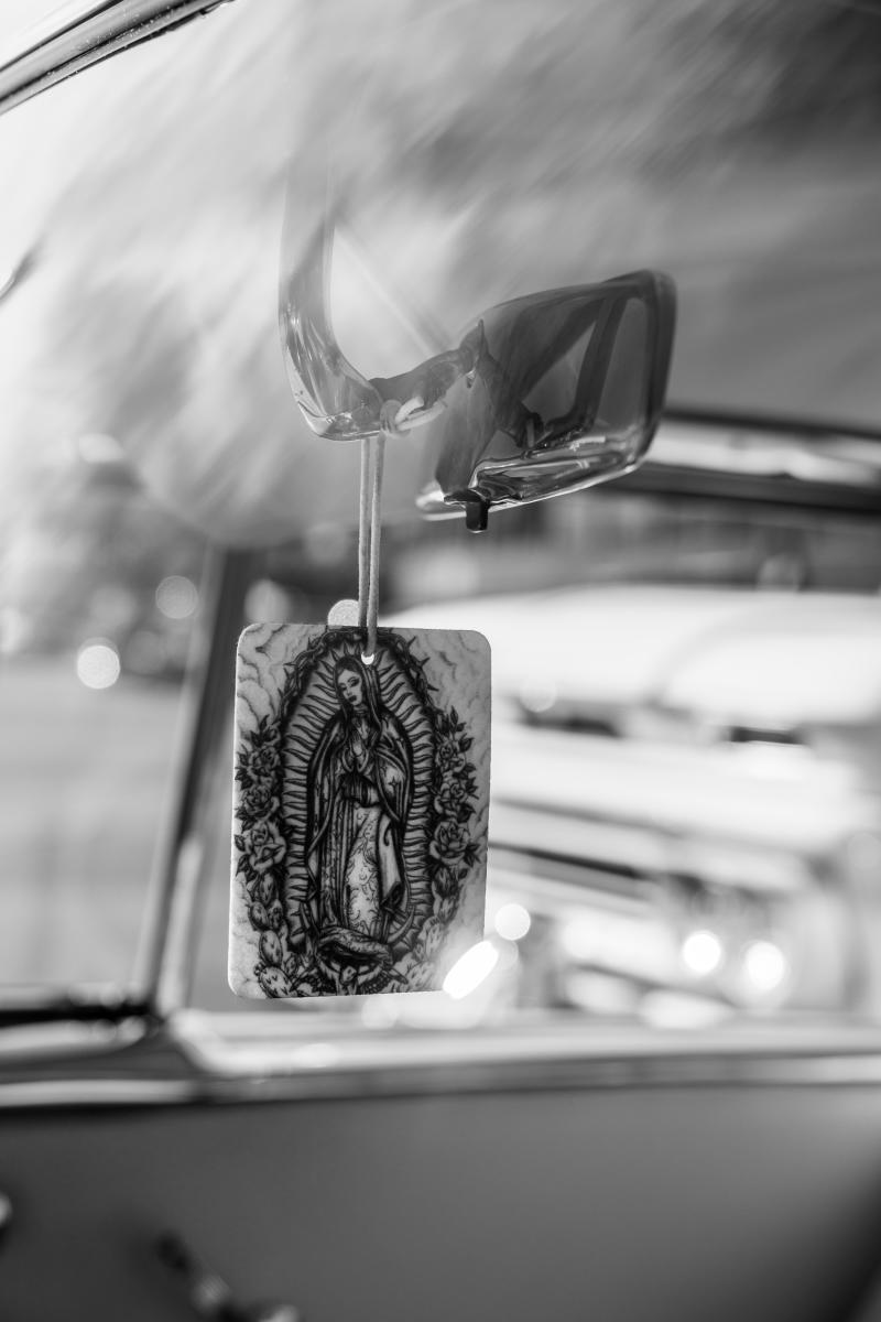 Black and white photo of a car interior with a religious card of the Virgin of Guadalupe hanging from the rearview mirror.