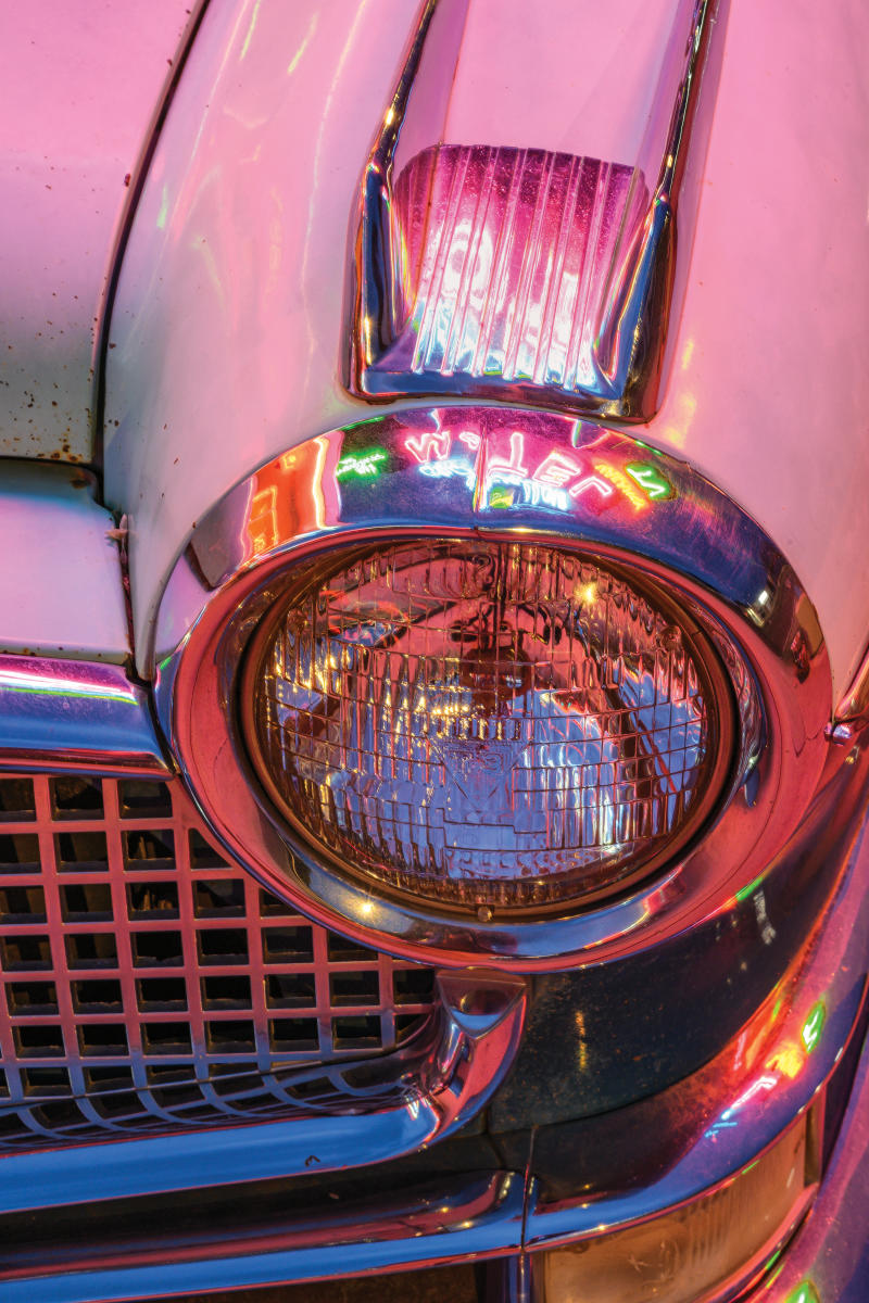 Close-up of a vintage car's headlight and chrome grille, bathed in vibrant neon pink and orange lights.
