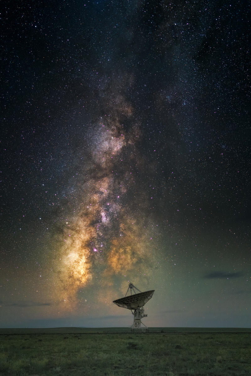 A large satellite dish sits in a grassy field, dwarfed by a brilliant Milky Way stretching across the night sky.