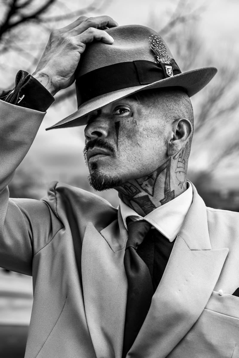 A sharply dressed man wearing a suit, vest, and matching hat stands outdoors in South San Jose Park, showing expressive eyes and visible tattoos.