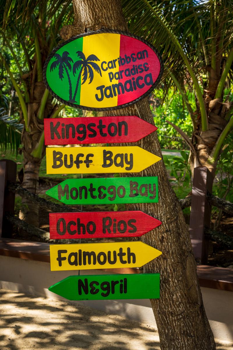 A wooden signpost with a colorful oval sign reading "Caribbean Paradise Jamaica" and arrows  pointing to different destinations in Jamaica: Kingston, Buff Bay, Montego Bay, Ocho Rios,  Falmouth, and Negril.
