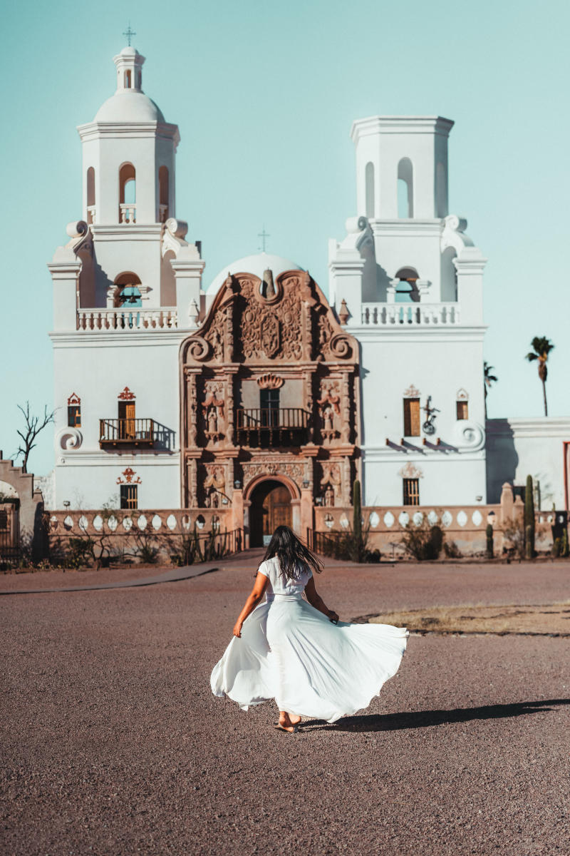 Woman in a white flowing dress walking towards the front of Mission San Xavier Del Bac, an ornately detailed Spanish, church-looking building