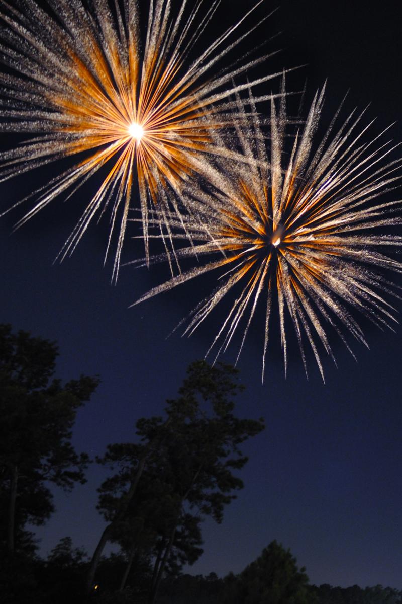 Orange fireworks explode over Lake Woodlands beside Lake Robbins Bridge and The Woodlands Towers.