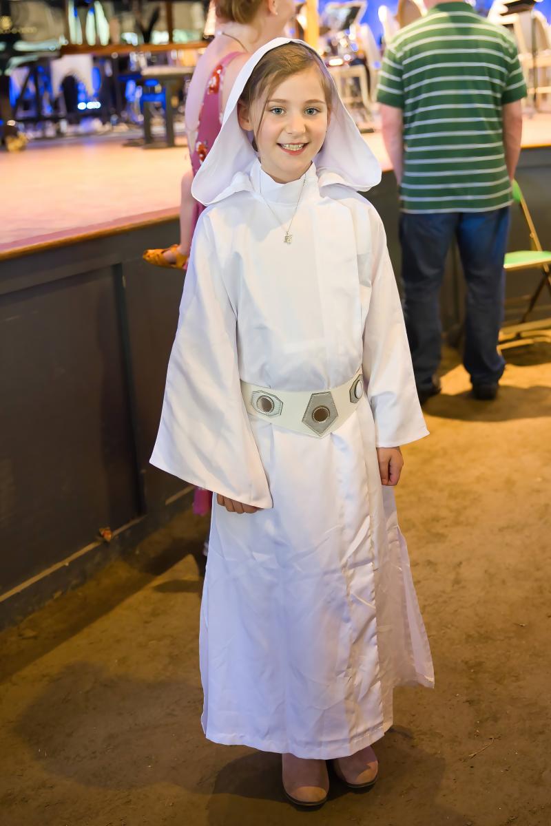A young white girl with blonde hair smiles at the camera. She is standing in front of a stage featuring a grand piano, a large golden harp, and chairs with music stands for an orchestra. The girl is wearing a costume based on Princess Leia from the Star Wars films: a white dress with a long skirt, wide sleeves, and a white hood resting halfway on her head. The costume also features a wide white belt with geometric metal accents, and she paired this look with a small gold necklace and pink suede boots.