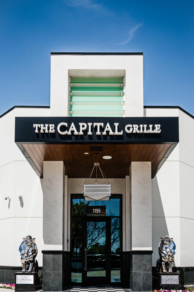 The front of The Capital Grille. It is a white, geometric building with black and silver accents. The window panels are a bright teal. Beside the front doors are a pair of silver metal lions.